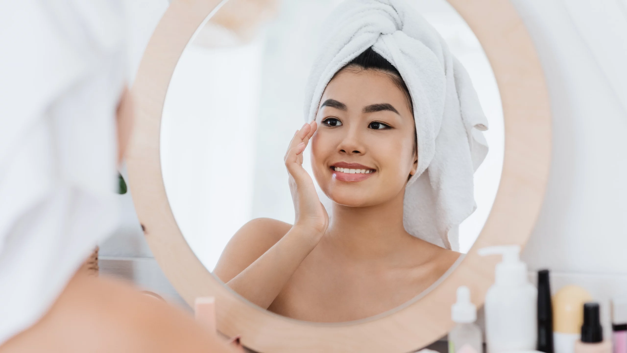 Asian woman looking in the mirror while wearing a towel over her head and touching her face.
