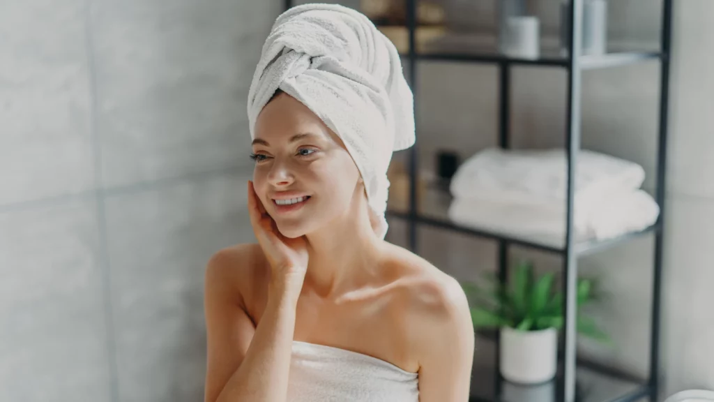 Woman wearing a bath towel and a towel over her hair touching her face and smiling.