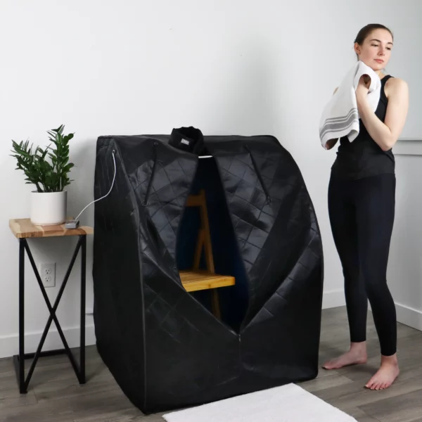 Woman holding a towel and standing beside a black UltraLux Infrared Foldaway Sauna.